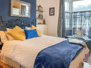 Una cama bien hecha con una colcha y almohadas estampadas, una mesita de noche con una lámpara, un espejo en la pared y una ventana con cortinas.