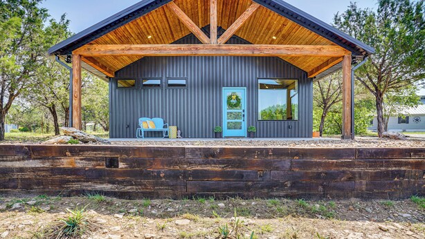A rustic cabin with a wooden roof and a blue door, surrounded by trees and a wooden deck. - Recently Built Cabin w/ Pavilion in Granbury! (Granbury)
