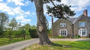 Une maison en pierre avec un grand arbre au premier plan, une allée pavée et une clôture en bois.