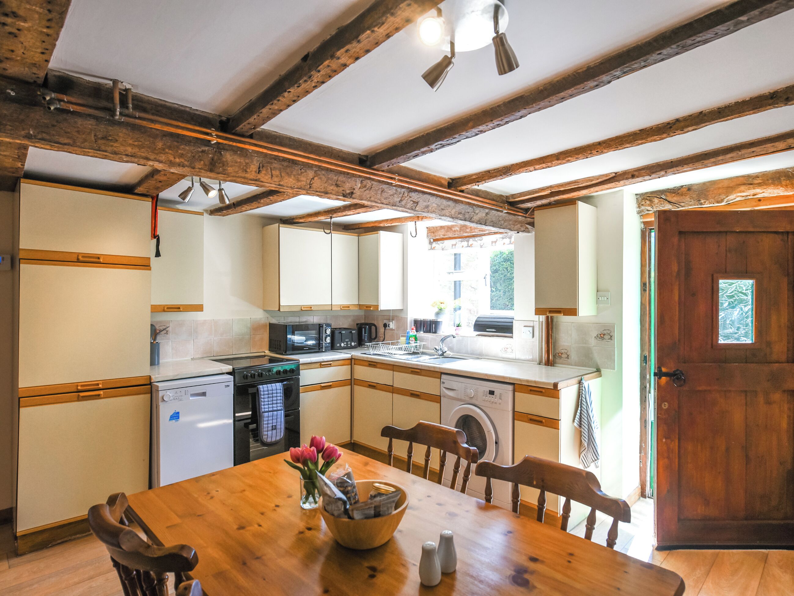 A kitchen with wooden beams, a dining table with chairs, a microwave, a coffee maker, and a washing machine.