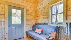 Ein Holzhütten-Interieur mit einer grauen Tür, einem blauen Sofa mit einem farbenfrohen Kissen, einem kleinen Holztisch mit einer Laterne und einem Fenster mit gemusterten Vorhängen.