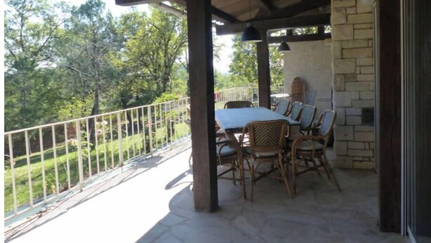 A covered patio with a table and chairs, a stone wall, and a view of trees. - Large campane house (Brignac-la-Plaine)