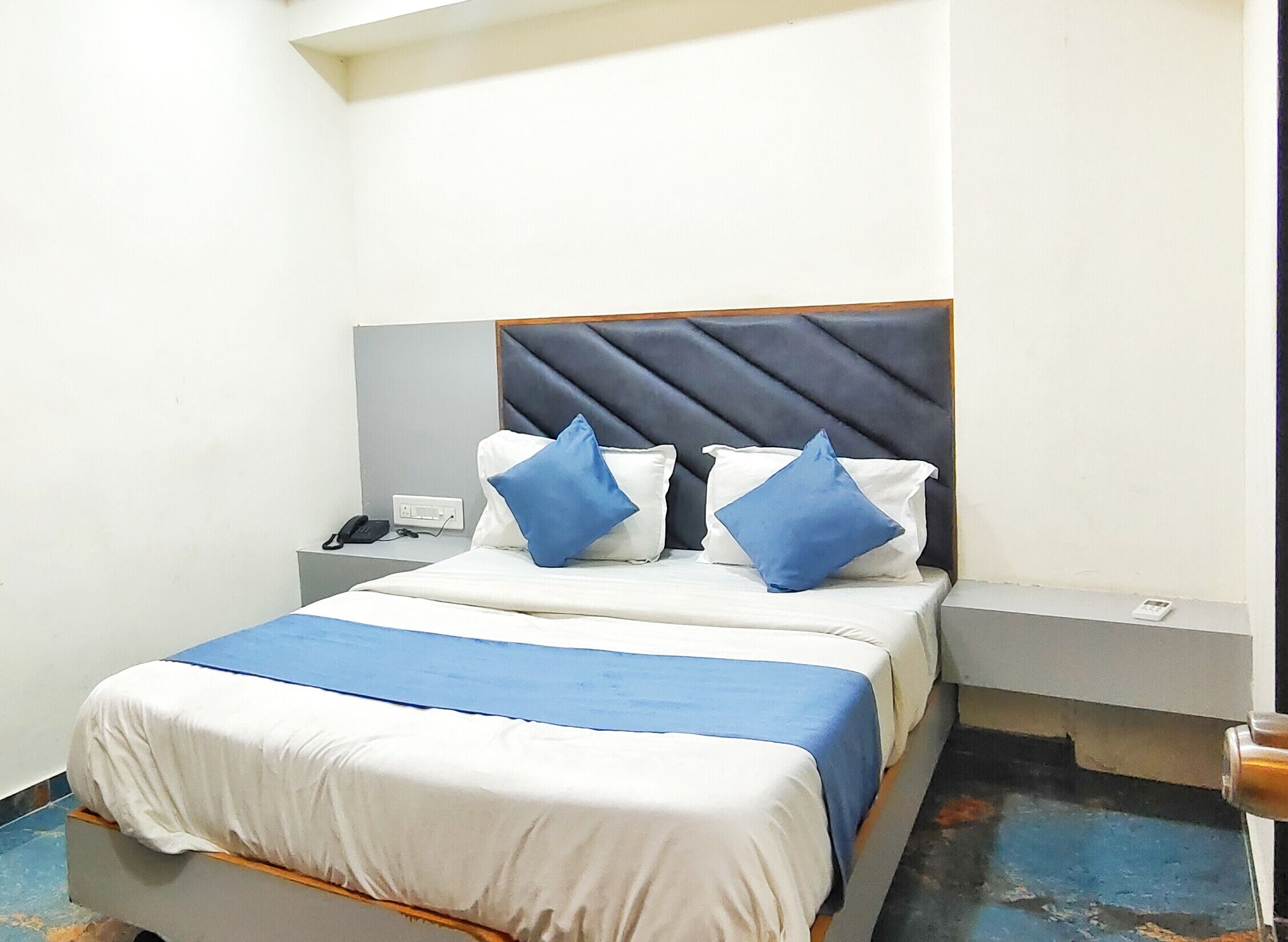 A neatly made bed with blue and white bedding, a headboard with a diamond pattern, and bedside tables with a phone and a remote control.