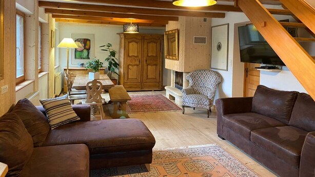 A cozy living room with a brown sofa, patterned rug, wooden beams, and a television.