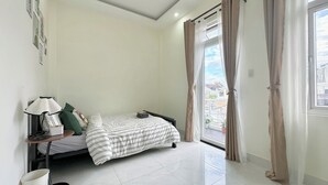 A bedroom with a bed, a bedside table with a lamp, a window with curtains, and a view of buildings outside. - Homestay Da Lat - The November Stay - Ngo Quyen (Cam Ly - Da Lat)