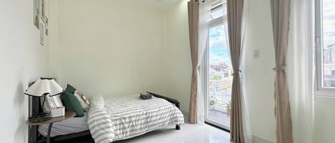 A bedroom with a bed, a bedside table with a lamp, a window with curtains, and a view of buildings outside.