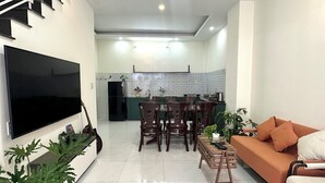 A modern living room with a flat-screen TV, a sofa, a dining area with chairs, and a kitchen in the background. - Homestay Da Lat - The November Stay - Ngo Quyen (Cam Ly - Da Lat)