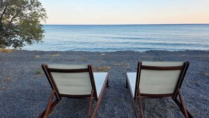 Two wooden lounge chairs with white cushions on a gravel beach with a view of the sea. - JM Dēos Suites (Viannos)