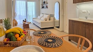 A modern living room with a sofa, a dining table with a fruit basket, and a kitchen area with wooden cabinets. - JM Dēos Suites (Viannos)