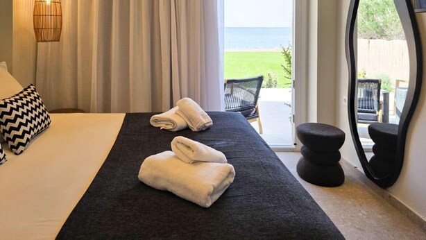 A hotel room with a bed, two towels, a chair, and a view of the outdoors. - JM Dēos Suites (Viannos)