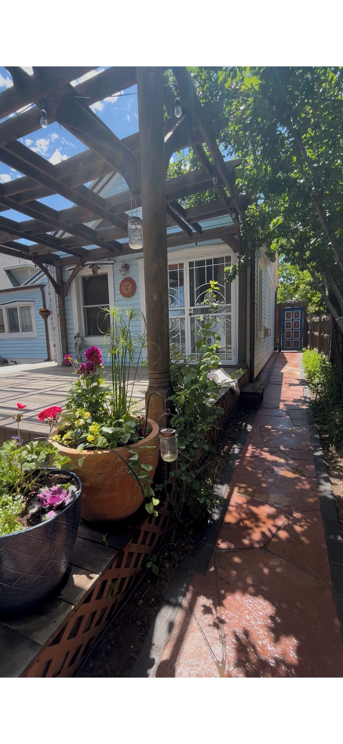 Un espace extérieur abrité, doté d’une pergola en bois, de plantes en pot et d’un chemin menant à une maison.