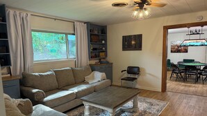 A cozy living room with a sofa, a coffee table, and a bookshelf. There is a window with curtains, a ceiling fan, and a painting on the wall. - Darbonne Ranch House - 12 beds! (Farmerville)