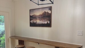 A room with a wooden bar counter, three stools, a hanging light fixture, and a landscape painting on the wall. - Darbonne Ranch House - 12 beds! (Farmerville)