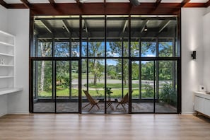 Une pièce avec de grandes fenêtres, un plancher en bois, une chaise, une petite table et une étagère.