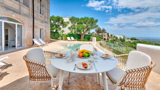A dining area with a table set for two, surrounded by wicker chairs, overlooking a pool and a scenic view of a coastal landscape.