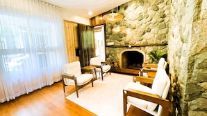 Una sala de estar con chimenea de piedra, muebles de madera y un ventanal con cortinas de gasa.