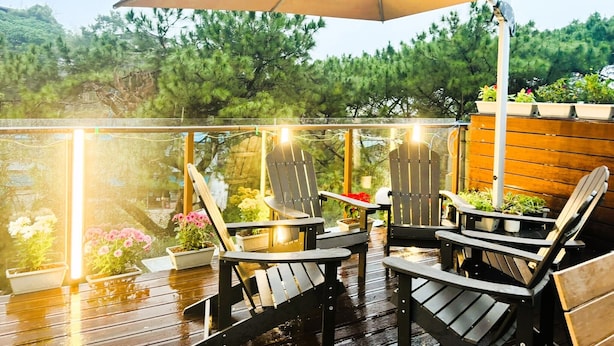 Una terraza al aire libre con sillas y mesas de madera, rodeada de plantas en macetas y un paraguas grande que ofrece sombra.