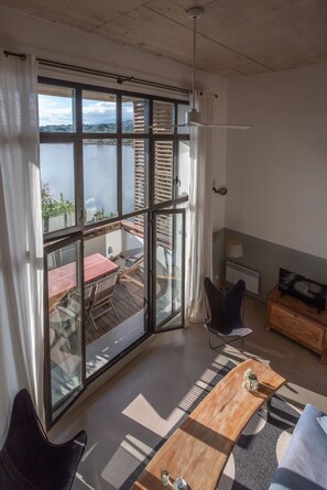 A spacious room with a large sliding glass door leading to a balcony overlooking a body of water. Inside, there is a wooden table, a sofa, and a chair. - Loft with a terrace overlooking the salt marshes of Porto-Vecchio (Porto-Vecchio)
