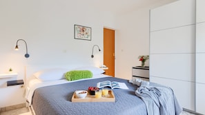 A modern bedroom with a bed, bedside lamps, a bedside table with a plant, a wooden nightstand, a bookshelf, and a door.