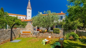 A courtyard with a stone wall, a wooden bench, and a lounge chair. A stone staircase leads to a building with a tower.