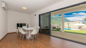 A room with a dining table and chairs, a flat-screen TV, and a sliding glass door leading to an outdoor area with a pool and mountains in the background.