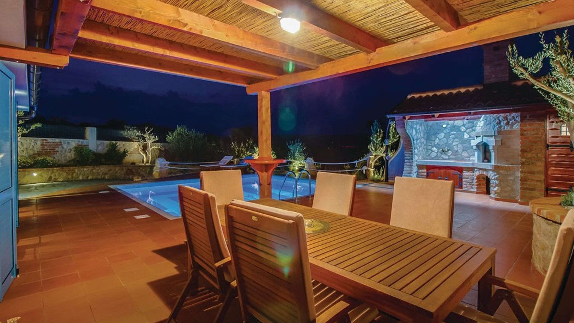 An outdoor patio with a pool, wooden furniture, and a stone fireplace.
