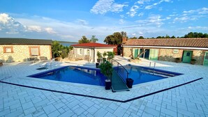 Une piscine entourée d’une terrasse carrelée, elle-même bordée de bâtiments en pierre aux toits rouges et aux volets verts.