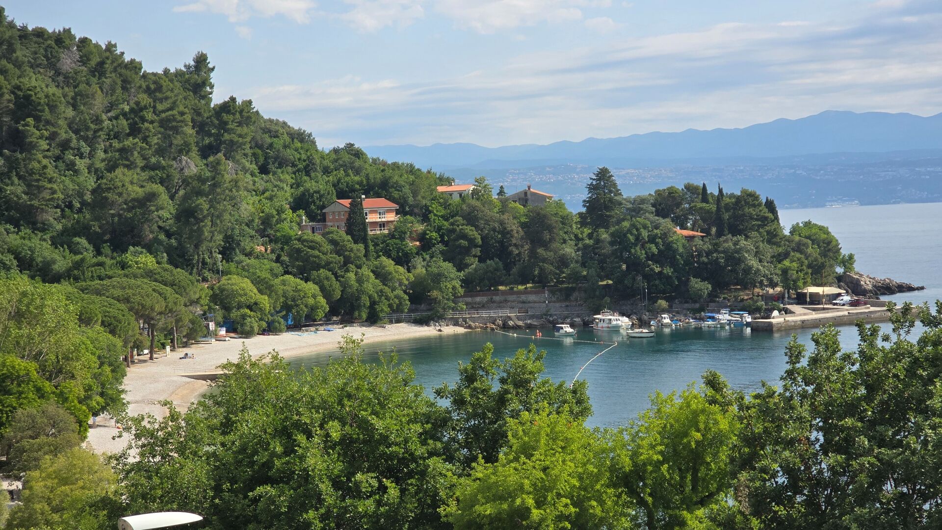 A coastal area with a beach, boats, and buildings surrounded by trees.