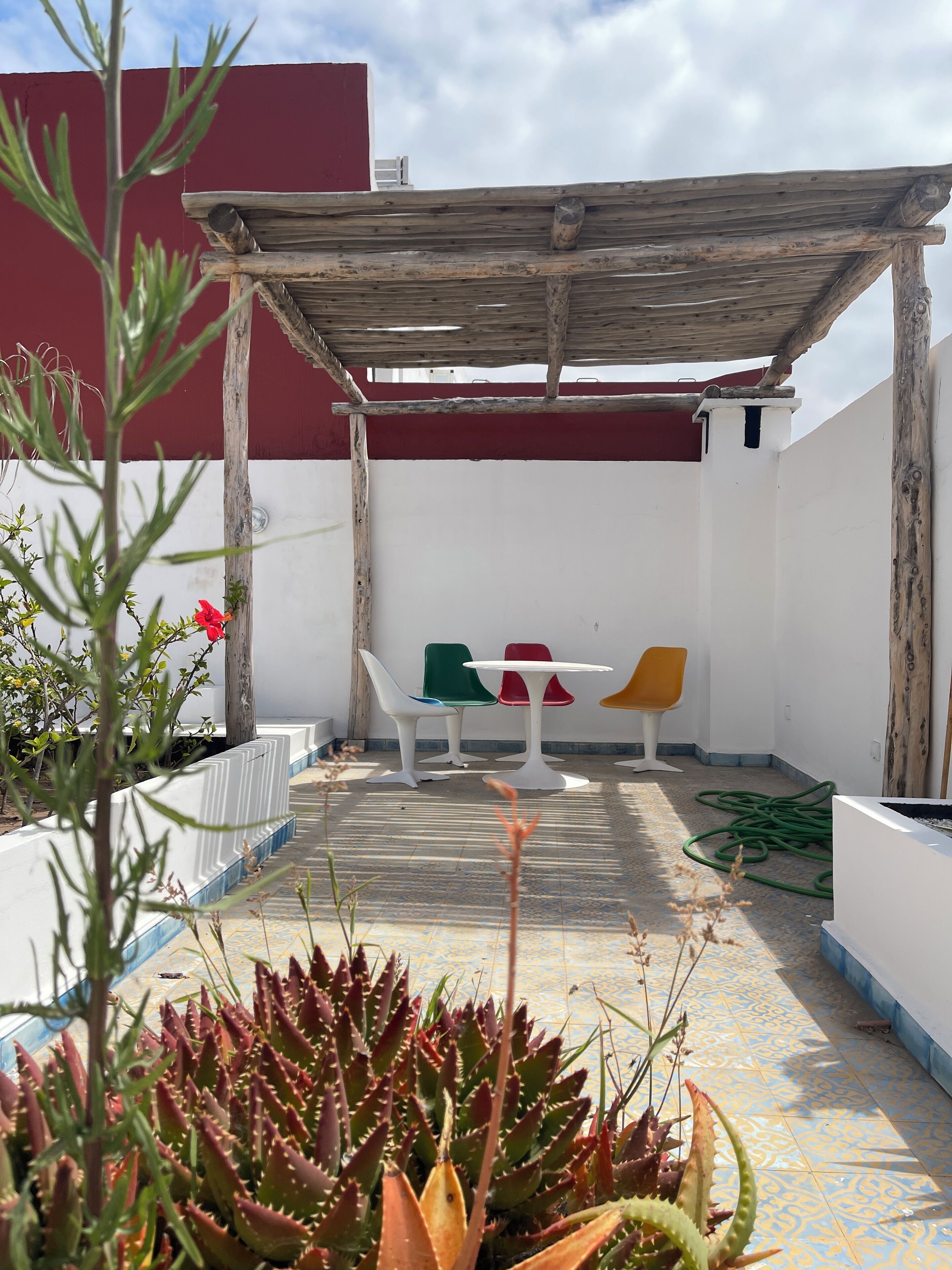 Un patio con una pérgola de madera, sillas blancas y coloridas, y una variedad de plantas, entre ellas suculentas.