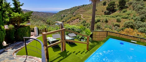 A swimming pool with a stone pathway leading to a seating area with sun loungers and a view of a hilly landscape.