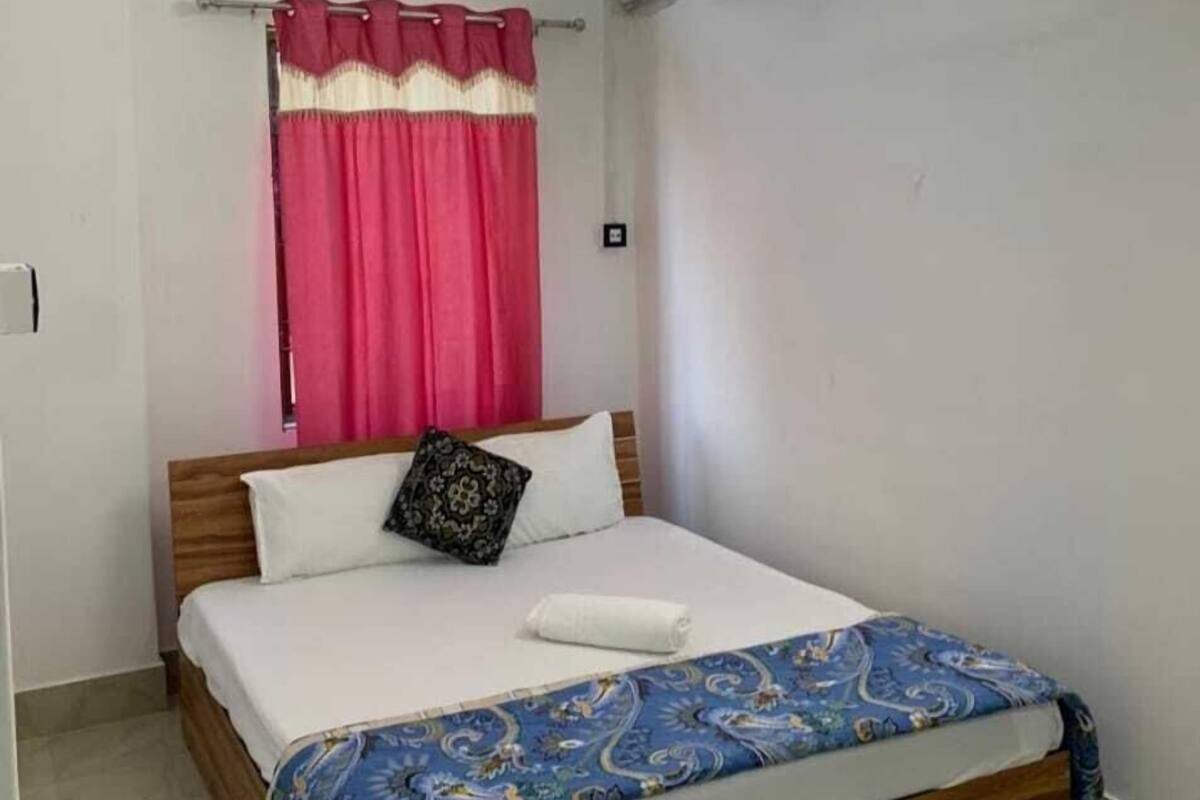 Ein Schlafzimmer mit einem hölzernen Bett, weißer Bettwäsche, einer gemusterten, blauen Decke und einem rosa Vorhang sowie einem Fenster mit Gitterstäben.