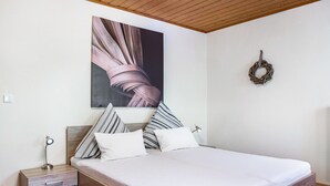 A bedroom with a wooden headboard, a bed with white bedding, striped pillows, a nightstand with a lamp, and a wall-mounted wreath.