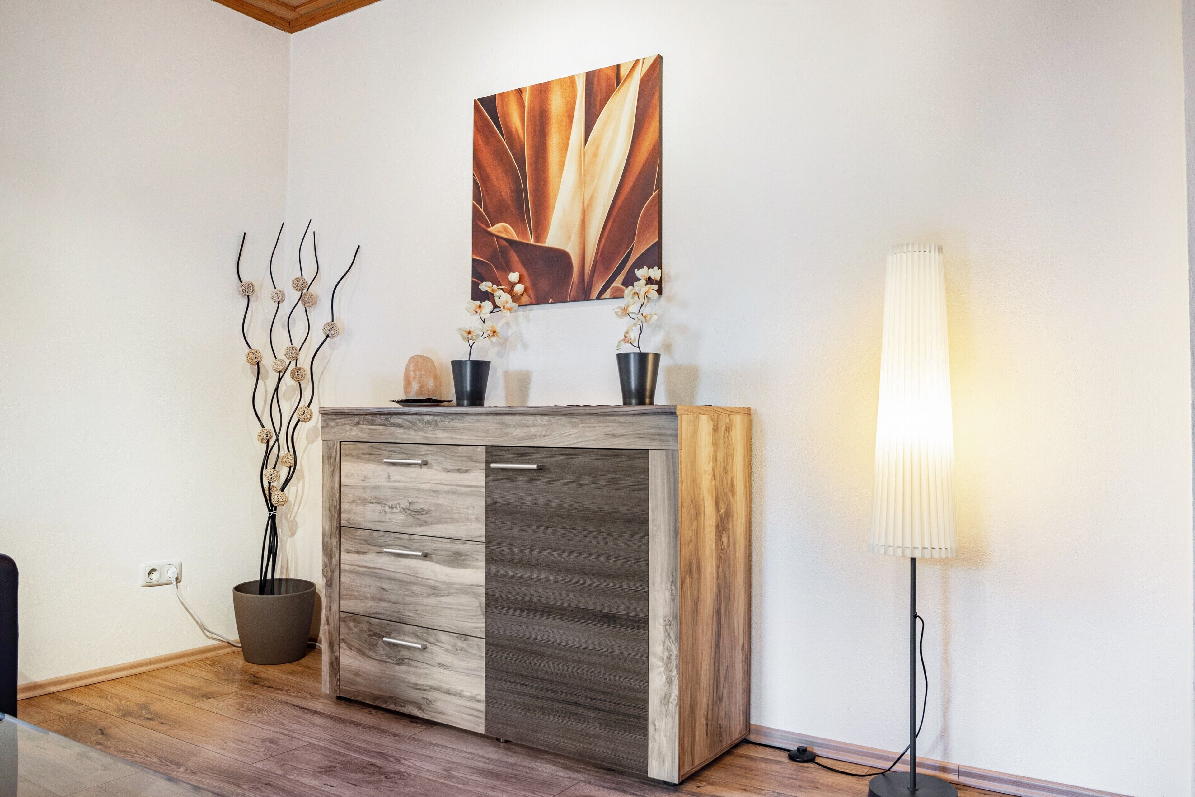 Una stanza con un armadio in legno, una pianta in vaso, una lampada da terra e un'opera d'arte appesa al muro.