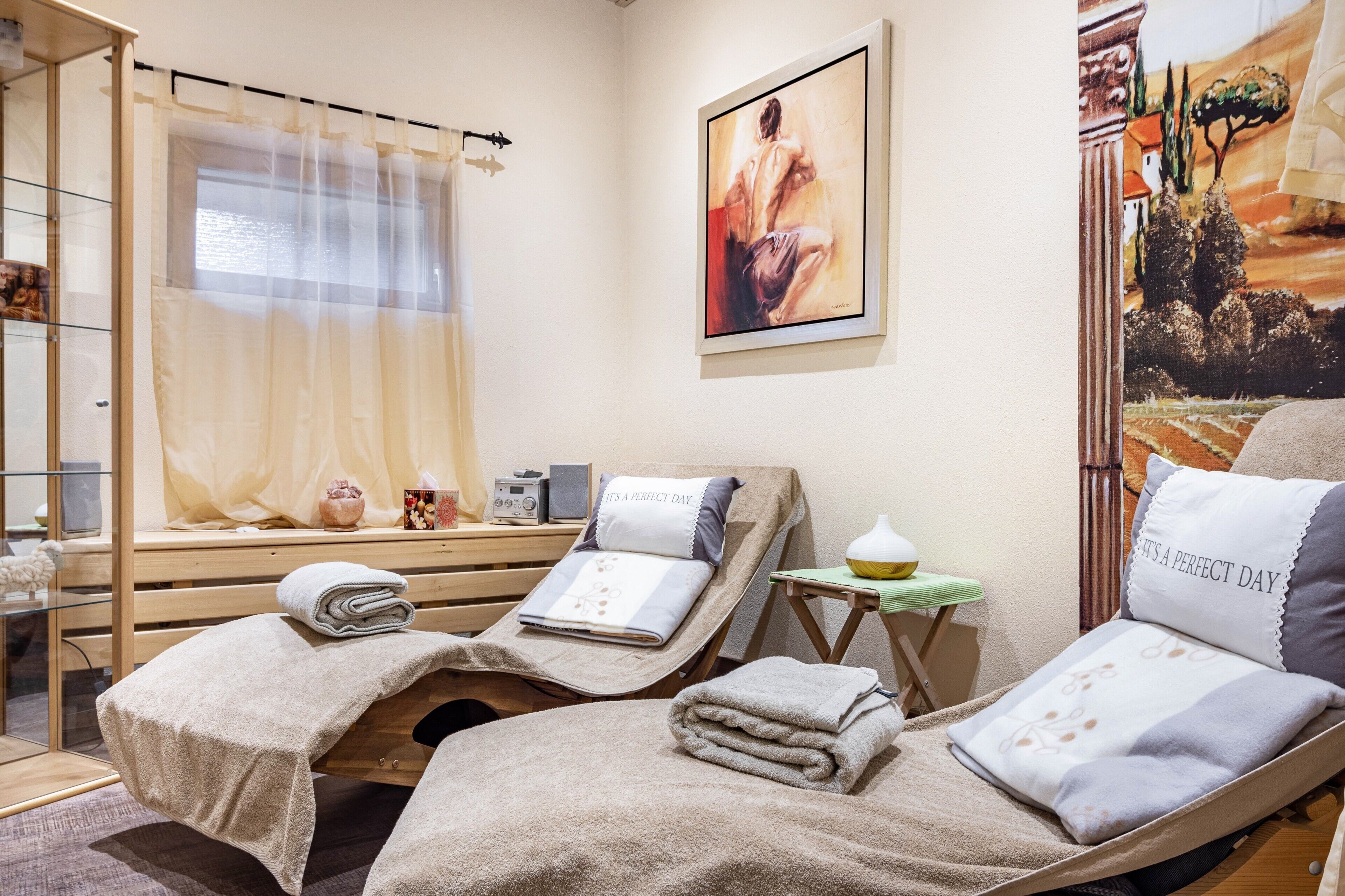 A spa room with two reclining chairs, a window with sheer curtains, a painting on the wall, and a small table with a lamp.