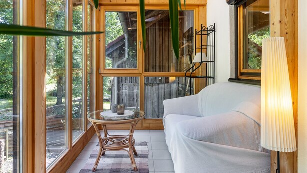 A wooden interior with large windows, a white sofa, a glass table, and a lamp.