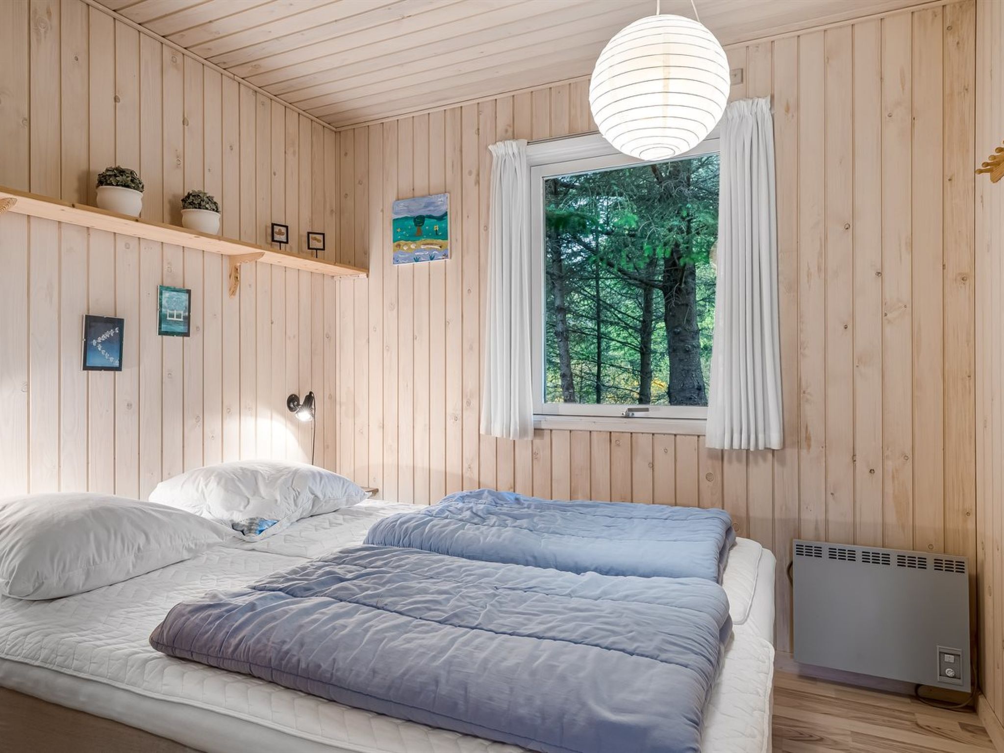 Ett sovrum med träpanel på väggarna, en säng, ett fönster med gardiner, ett element och vägghängda hyllor med krukväxter.