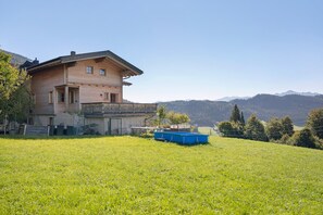 Ein Holzhäuschen mit Balkon, umgeben von einer grünen Wiese und Bergen im Hintergrund.
