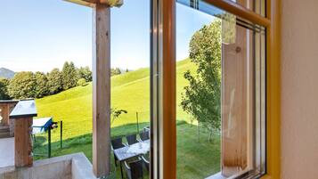 Blick durch ein Fenster auf eine Holzterrasse mit Tisch und Stühlen, einen Rasen und einen Baum.