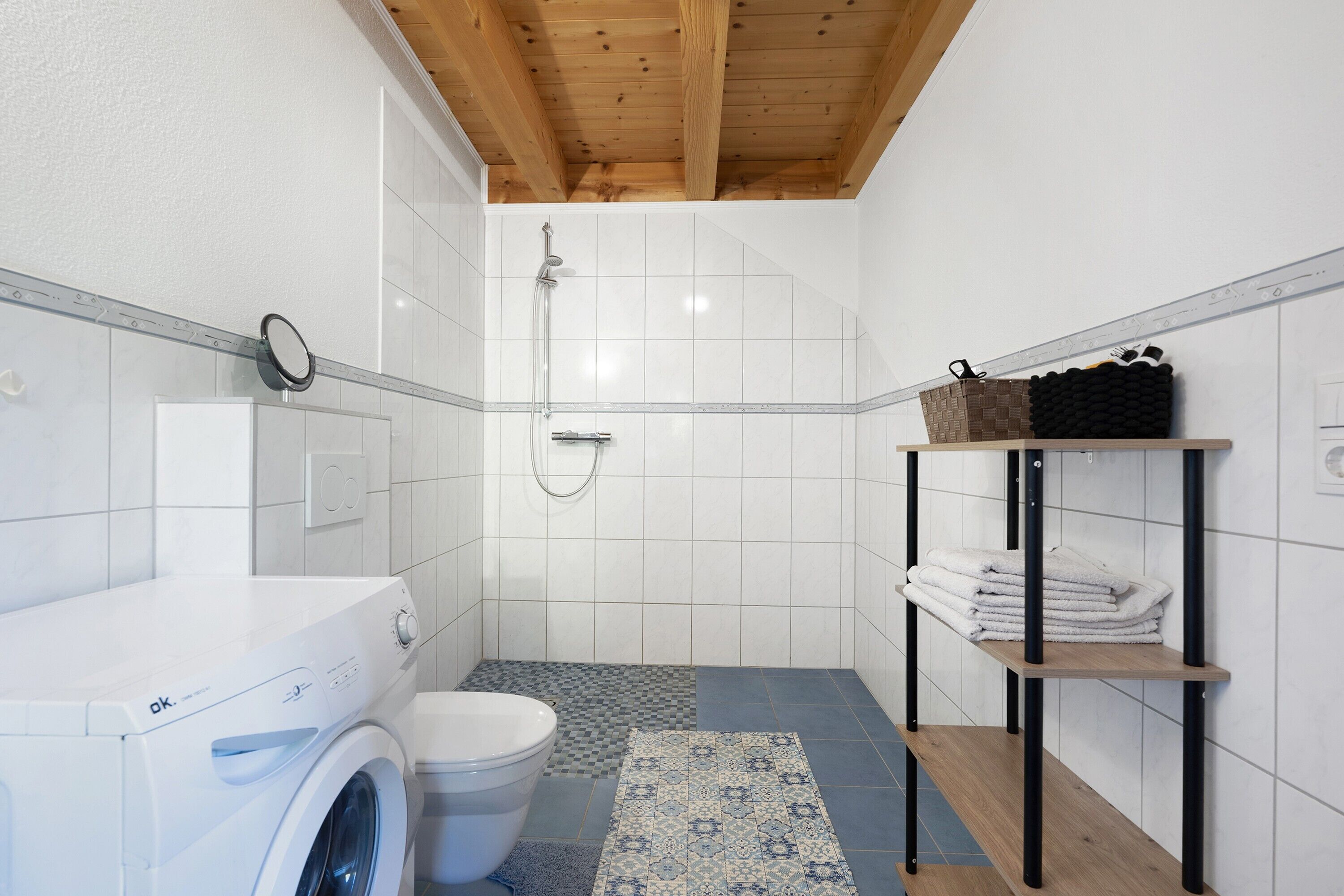 Une petite salle de bain équipée d’un lave-linge, de toilettes, d’une douche et d’une étagère où sont rangées les serviettes.