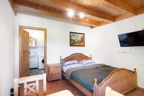 Ein Schlafzimmer mit einem hölzernen Bett, einem an der Wand montierten Fernseher, einem Gemälde über dem Bett und einem Waschbereich, der durch eine geöffnete Tür sichtbar ist.