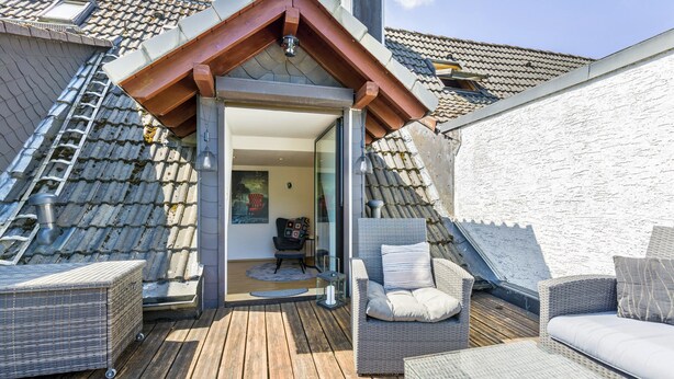 Una terraza de madera con mobiliario de mimbre, una puerta de cristal y techo de tejas.