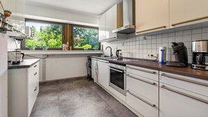 Una cocina moderna con armarios blancos, una campana extractora de acero inoxidable, horno empotrado, cafetera y ventana con vista a zonas verdes.