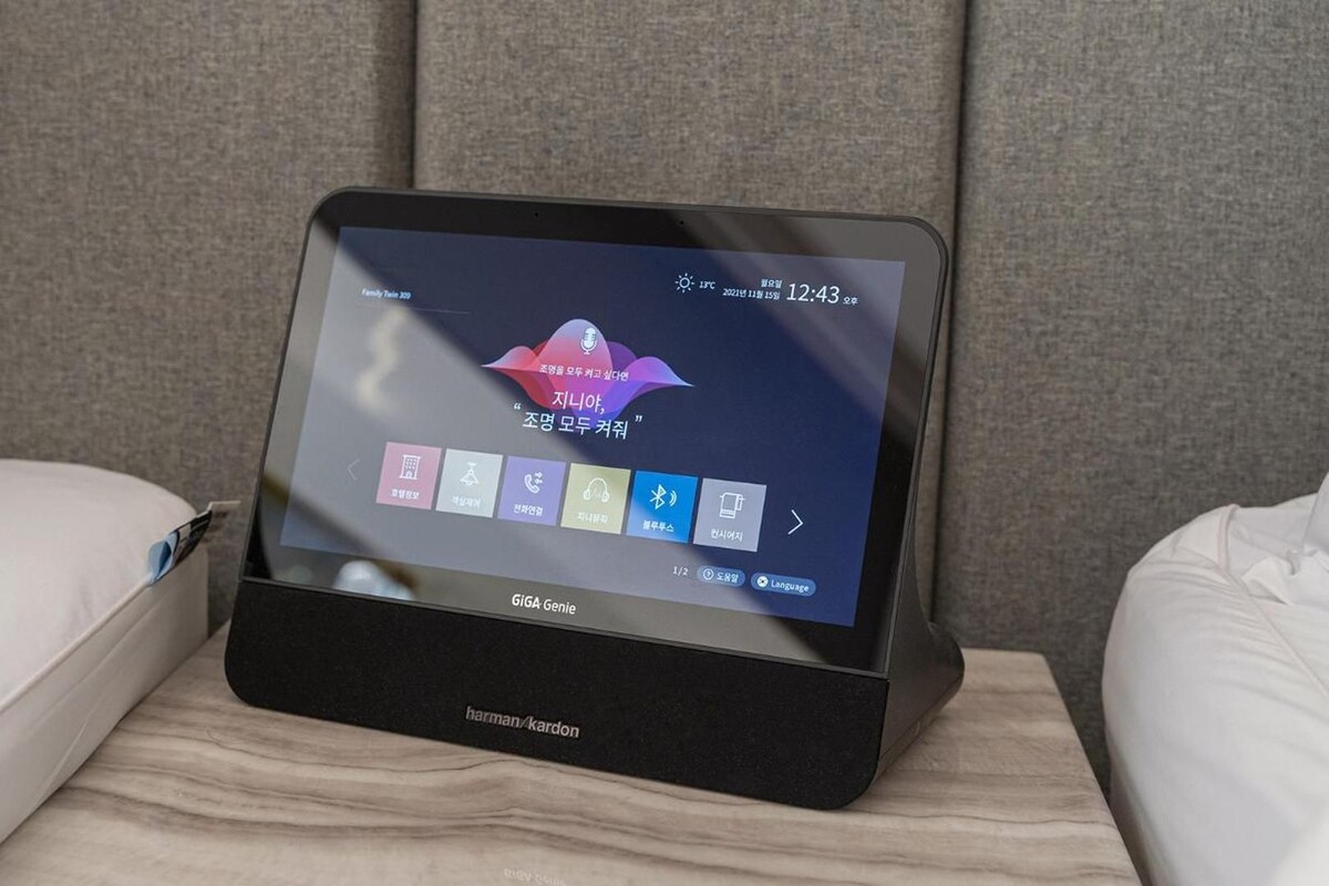 A hotel room with a bed, a bedside table, and a black electronic device displaying a weather app.