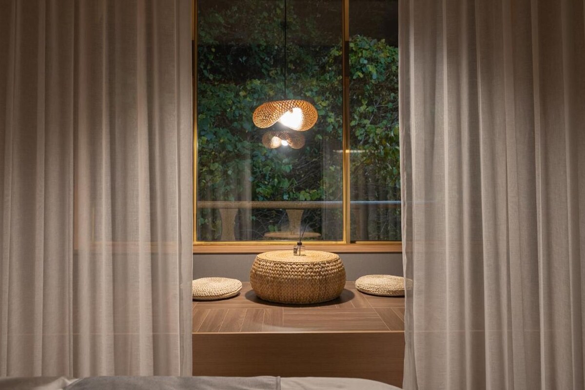 A room with a window, curtains, a woven lamp, and a wooden bench.