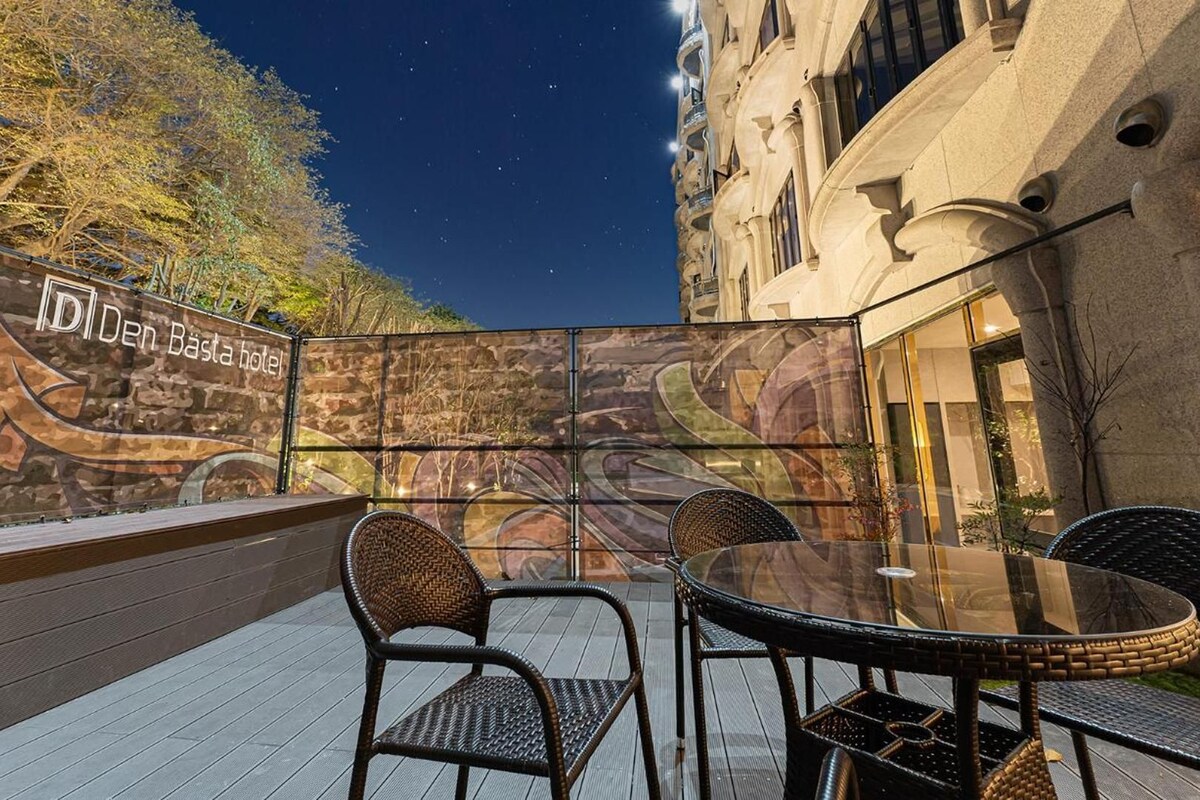 An outdoor seating area with wicker chairs and a glass table, a wall with a mural, and a building with classical architecture.