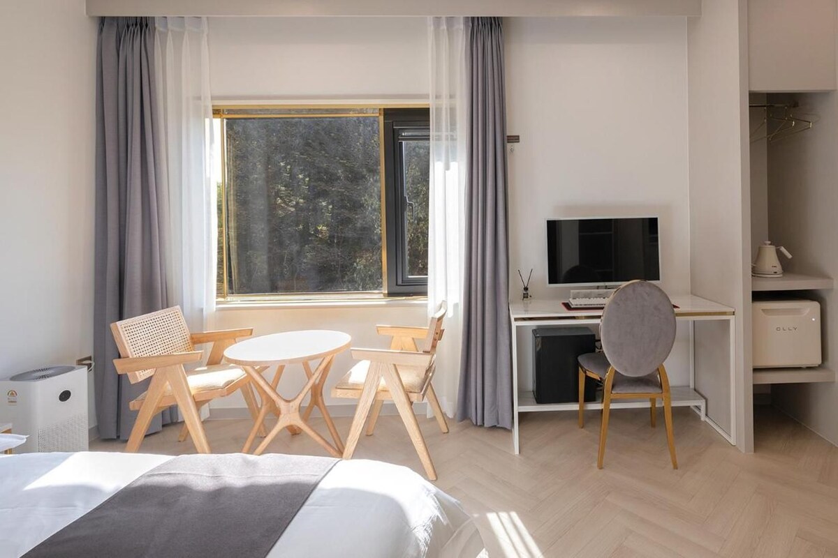 A modern hotel room with a bed, a desk with a computer, a chair, and a view of trees through the window.