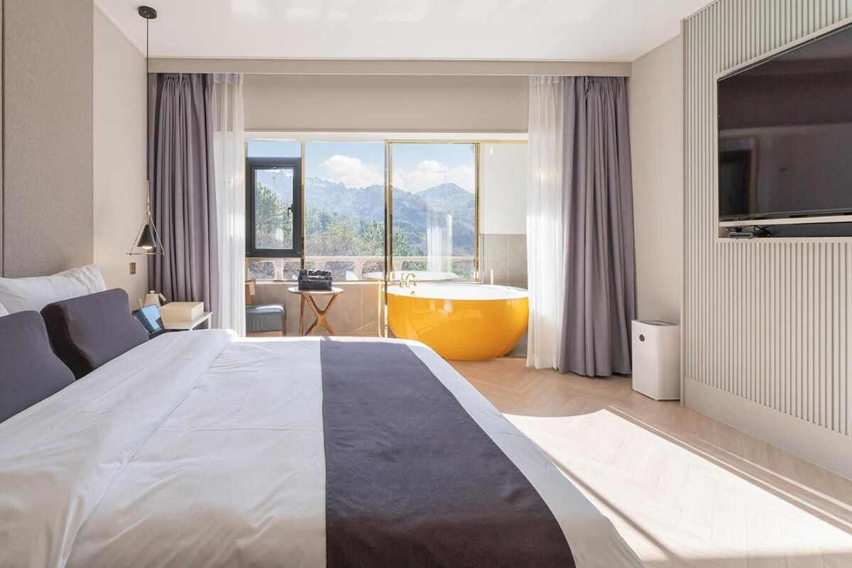 A hotel room with a large bed, a view of mountains, a bathtub, and a desk with a chair.
