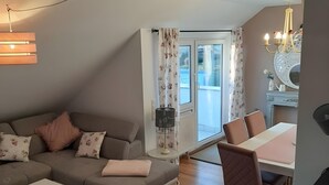 A living room with a sofa, a dining table, and chairs. There is a window with curtains, a chandelier, and a radiator.