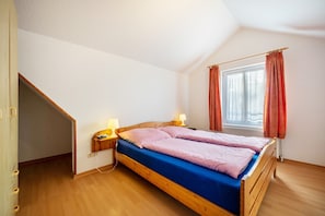 Une chambre avec un plafond en pente, un lit avec un matelas bleu, une tête de lit en bois, deux lampes de chevet et une fenêtre avec des rideaux.