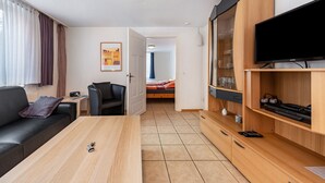 Una habitación de hotel moderna con sofá, una mesita de centro de madera, un televisor sobre un mueble de madera, un escritorio con silla, y una cama con cortina azul.